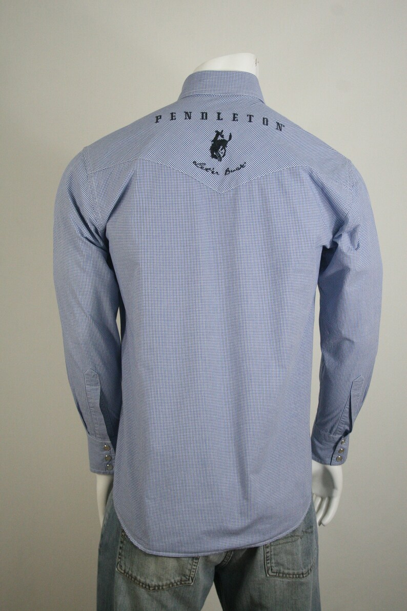 May include: A blue and white gingham long-sleeve shirt with a black embroidered logo on the back that says "PENDLETON" and features a rider on a horse. The logo also includes the text "Western Wear".