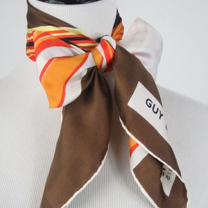 May include: A brown silk scarf with a white border and a colorful striped pattern. The scarf is tied around a mannequin's neck and has a white label with the text "GUY LAROCHE" printed on it.