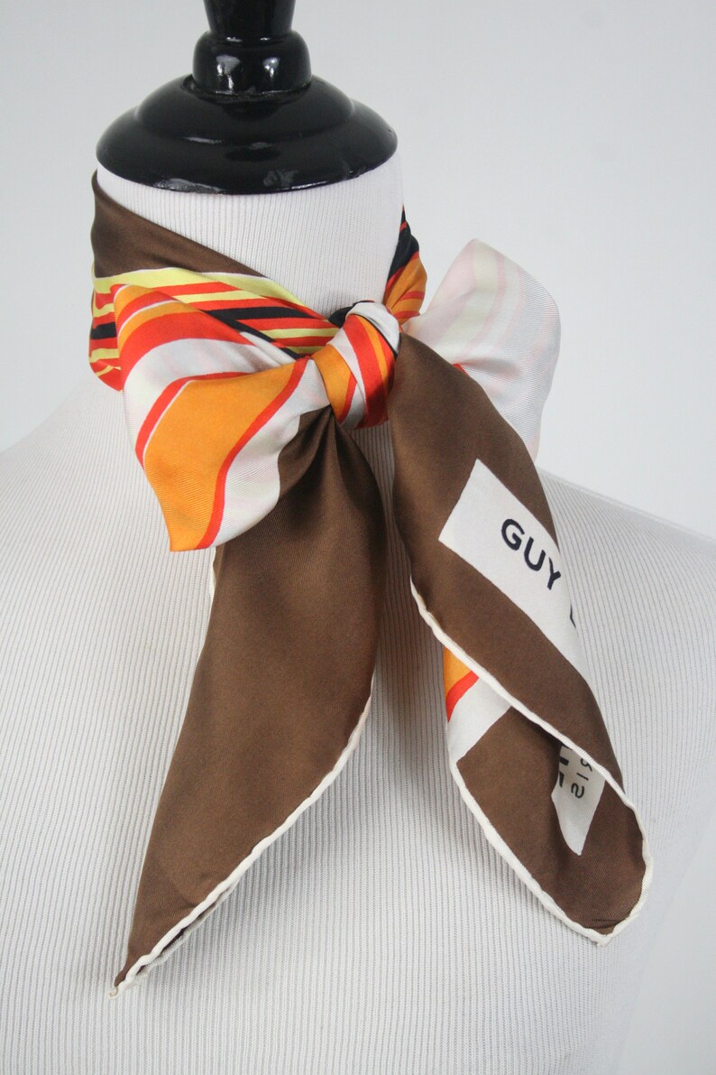 May include: A brown silk scarf with a white border and a colorful striped pattern. The scarf is tied around a mannequin's neck and has a white label with the text "GUY LAROCHE" printed on it.
