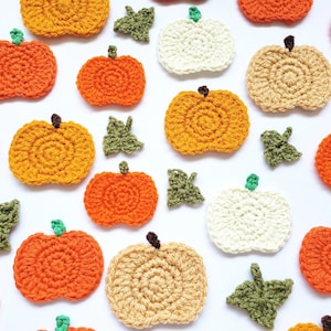 May include: A collection of crocheted pumpkins in various colours, including orange, yellow, and white, with green crocheted leaves scattered around them.