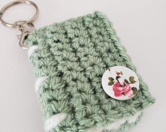 Puede incluir: Un llavero hecho a mano, tejido a ganchillo, en color verde suave con detalles blancos. El llavero presenta un botón floral decorativo y un llavero y cierre plateados. El artículo está diseñado para guardar llaves o objetos pequeños.