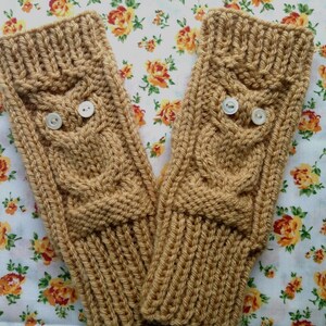 Puede incluir: Un par de guantes sin dedos de color beige con un diseño de búho en punto de ochos. Cada guante tiene dos pequeños botones blancos que sirven como ojos para el motivo del búho. Los guantes tienen un puño acanalado en la parte superior e inferior.
