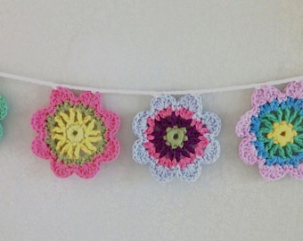May include: A decorative garland featuring five multi-colored crocheted flowers strung along a white cord. Each flower displays a unique combination of vibrant yarn colors including pink, yellow, blue, green, purple, and orange in a circular pattern.