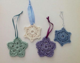 May include: Four crocheted star-shaped ornaments in light green, cream, purple, and blue. Each star features a central hole and is attached to a hanging ribbon: a teal satin ribbon, a blue and white gingham ribbon, a purple ribbon, and a silver cord.