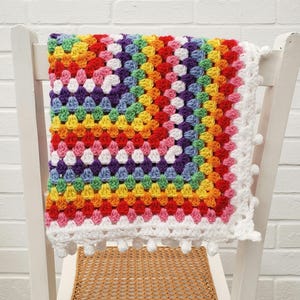 May include: A rainbow granny square crochet blanket with a white border and pom-poms. The blanket is draped over a white chair.