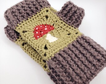 May include: A pair of fingerless gloves crocheted with dark brown yarn at the cuffs and thumb opening. The center features a square motif in olive green yarn with a red and white mushroom graphic embroidered in the middle of the square pattern.