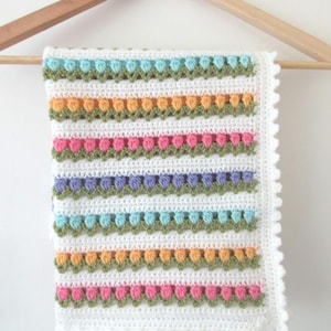 May include: A white crochet blanket with horizontal stripes of colorful floral patterns in pink, orange, blue, and green. The blanket is hanging on a wooden hanger, showcasing its intricate design and scalloped edges. The blanket is approximately 30 inches wide and 36 inches long.
