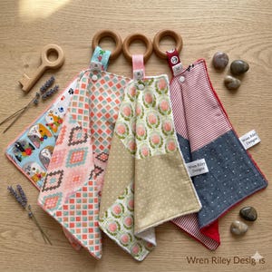May include: A collection of colorful baby teething toys with wooden rings and patterned fabric. Designs include cats, geometric shapes, and floral patterns. The toys are in shades of pink, blue, and beige, with red and white accents. The wooden key is also visible.