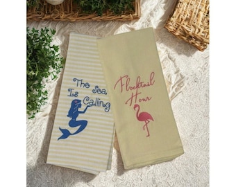 May include: Two decorative kitchen towels. One towel is cream-colored with yellow stripes and the text "The Sea Is Calling" and a blue mermaid graphic. The other towel is light yellow with the text "Flocktail Hour" and a pink flamingo graphic.