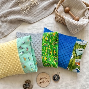 Könnte beinhalten: Eine Auswahl an Babykissen in Gelb, Grau, Grün und Blau mit verschiedenen Mustern. Einige Kissen haben strukturierte Oberflächen und Prints mit Fahrzeugen oder Campingmotiven. Ein Holzschild zeigt den Text "Travel Buddies".