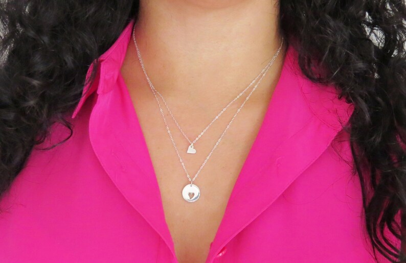 May include: A silver necklace with two pendants. The top pendant is a small heart, and the bottom pendant is a larger circle with a heart cut out of the center.