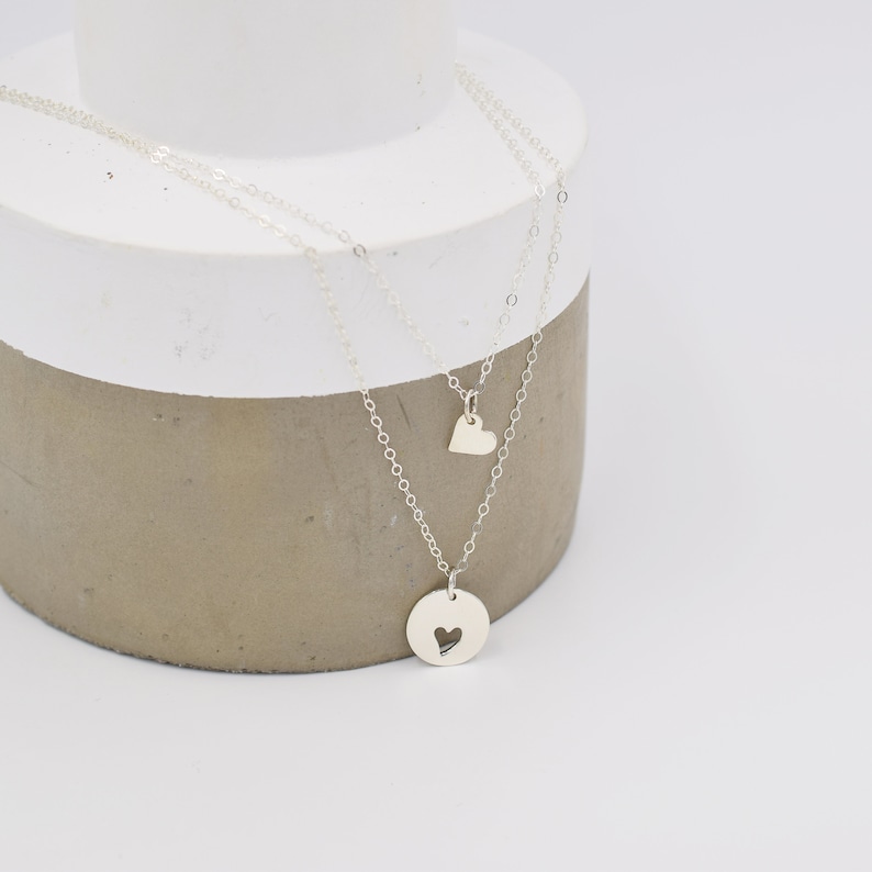 May include: A silver necklace with two heart-shaped pendants. The larger pendant has a heart-shaped cutout. The necklace is layered on a gray surface.