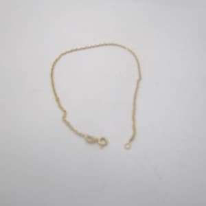 May include: A delicate gold-colored chain bracelet with a small clasp and a ring on the end. The bracelet is lying on a white surface. The chain appears to be made of small, twisted links.