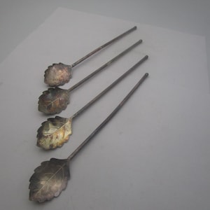 May include: Four silver-toned spoons with leaf-shaped bowls and long, slender handles. The spoons are arranged diagonally, showcasing their intricate leaf design. The spoons appear to be vintage or antique, adding a touch of elegance.