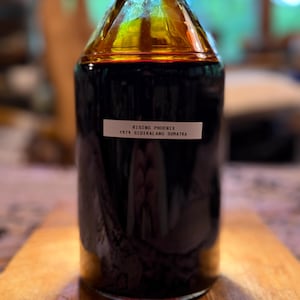Puede incluir: Una botella de vidrio transparente llena de un líquido ámbar oscuro, con una tapa azul. Una etiqueta blanca en la botella dice "RISING PHOENIX 1979 SIDIKALANG SUMATRA". La botella está sobre una bandeja de madera.