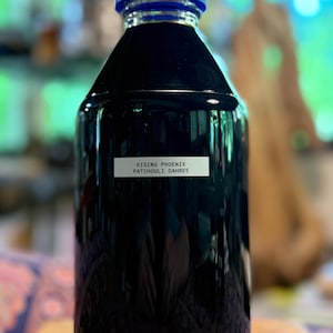 May include: A glass bottle containing a dark liquid, with a blue cap. The label on the bottle reads "RISING PHOENIX PATCHOULI DAHREE". The bottle is on a wooden surface. The background is blurred.