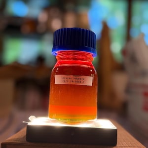 May include: A small glass bottle with a blue cap and a label that reads "RISING-PHOENIX 2025-TRIPURA-1." The bottle contains a layered liquid with red, orange, and yellow hues. It sits on a small, illuminated platform.