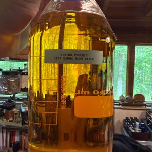 May include: A clear glass bottle with a blue cap, filled with a golden-yellow liquid. The bottle has a label that reads "RISING PHOENIX 2024 BANDA ACEH PATCH". The bottle is held by a person.