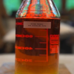 Può includere: Una bottiglia di vetro trasparente riempita con un liquido a strati, che passa dal giallo in basso all'ambra e al rosso in alto. L'etichetta recita "RISING PHOENIX 2010 SIDIKALANG PATCH". Sono presenti segni rossi per le misurazioni del volume.