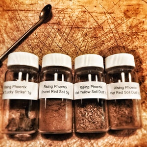 May include: Four small glass jars with white lids, each labeled "Rising Phoenix" with different soil types: "Lucky Strike," "Brunei Red Soil," "Viet Yellow Soil Dust," and "Viet Red Soil Dust." A small spoon rests above the jars. The background is a wooden surface.