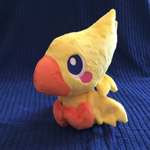 May include: Yellow and orange plush toy of a bird-like creature with large eyes and a beak. The toy is sitting on a blue surface.