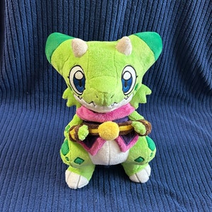 May include: Green plush toy with a pink and purple scarf, holding a gold and brown scroll. The toy has large blue eyes and a toothy grin.
