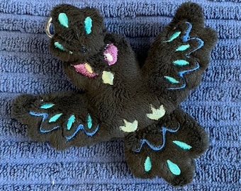 Puede incluir: Pájaro de peluche negro con bordados azules y turquesa. El pájaro tiene un pequeño anillo sujeto a su cabeza.