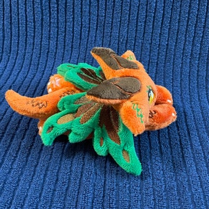 Puede incluir: Un peque&ntilde;o drag&oacute;n de peluche naranja y verde con detalles marrones. El drag&oacute;n tiene alas grandes de color verde y una peque&ntilde;a nariz marr&oacute;n.