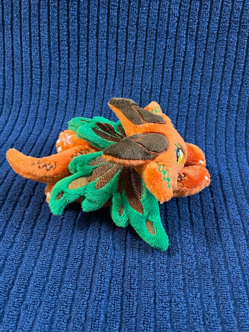 Puede incluir: Un peque&ntilde;o drag&oacute;n de peluche naranja y verde con detalles marrones. El drag&oacute;n tiene alas grandes de color verde y una peque&ntilde;a nariz marr&oacute;n.