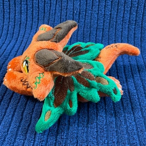 Puede incluir: Un drag&oacute;n de peluche naranja y verde con detalles marrones. El drag&oacute;n tiene un ala verde grande y una cola peque&ntilde;a.