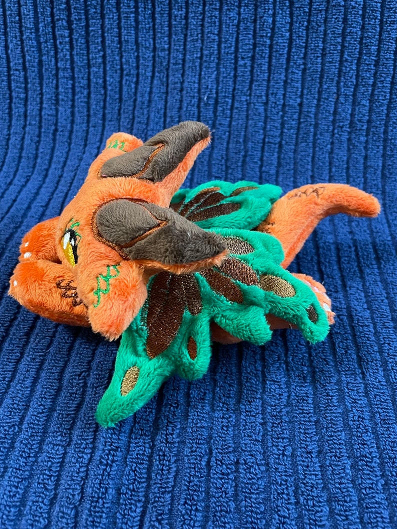 Puede incluir: Un drag&oacute;n de peluche naranja y verde con detalles marrones. El drag&oacute;n tiene un ala verde grande y una cola peque&ntilde;a.