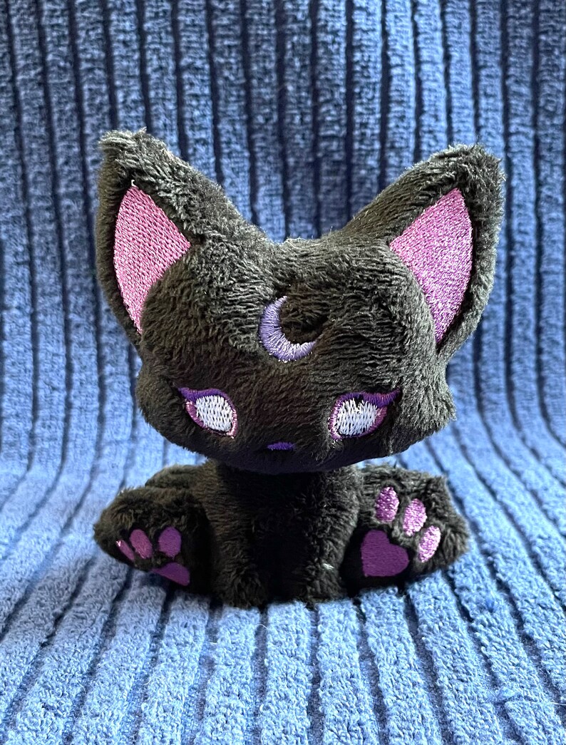 May include: A black plush cat with pink ears and paws. The cat has a crescent moon embroidered on its forehead and white eyes with purple eyelashes.