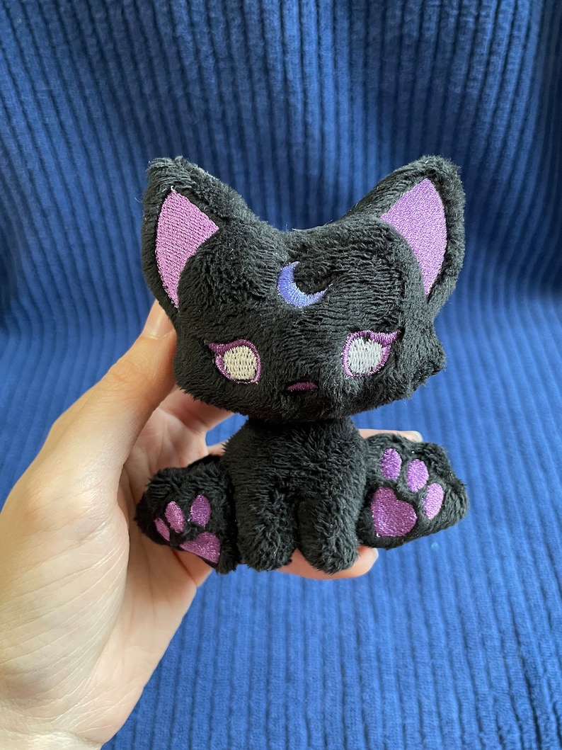 May include: Black plush cat toy with purple ears, paws, and a crescent moon on its forehead. The cat has white eyes with purple eyelashes.