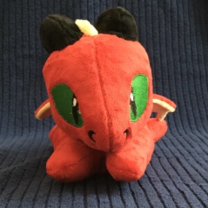 May include: A plush red dragon toy with black ears, green eyes, and small wings. The dragon is sitting on a blue textured surface. The toy is soft and cuddly, perfect for children.