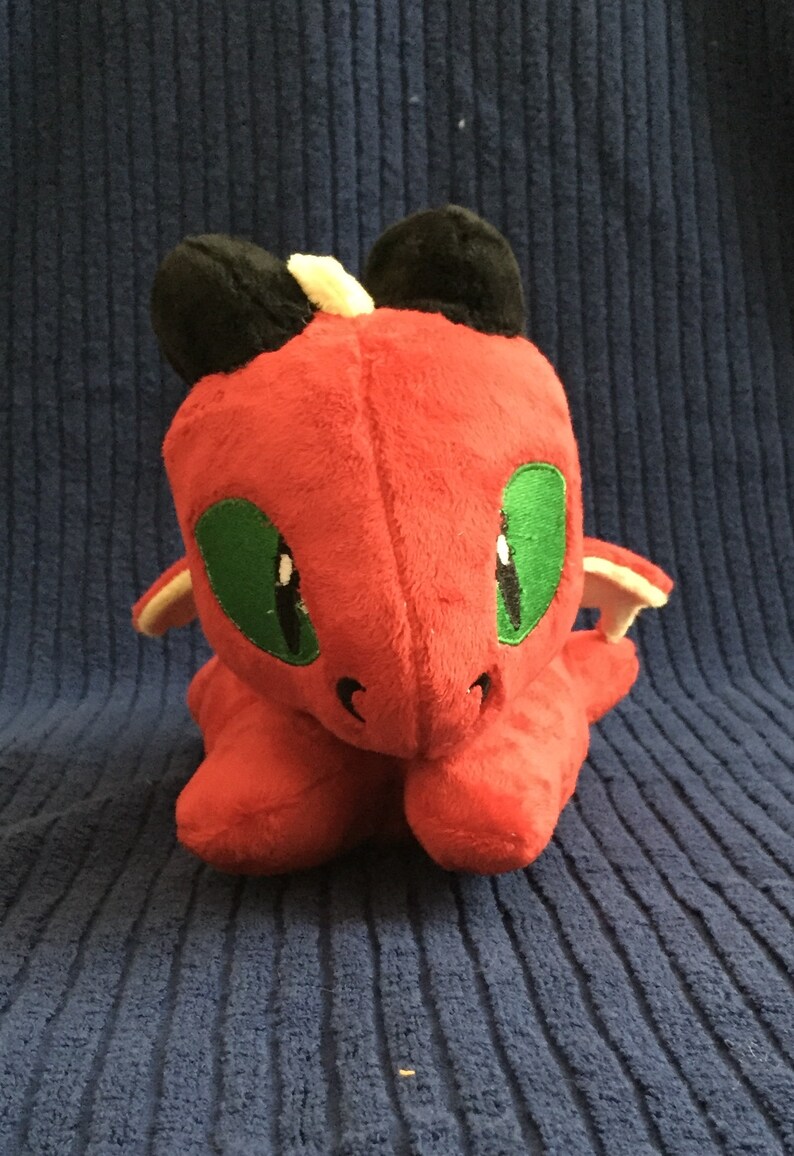 May include: A plush red dragon toy with black ears, green eyes, and small wings. The dragon is sitting on a blue textured surface. The toy is soft and cuddly, perfect for children.