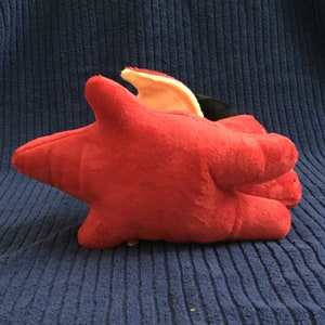 May include: A plush, red star-shaped toy with a yellow and black accent. The toy is made of soft material and is resting on a blue textured surface. The toy has a unique shape.