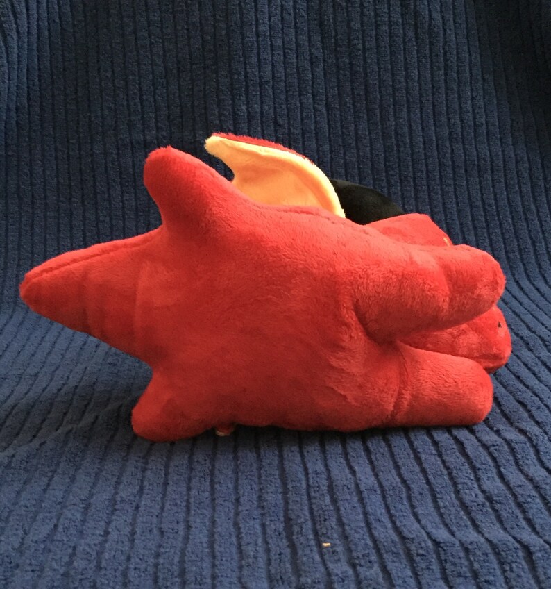 May include: A plush, red star-shaped toy with a yellow and black accent. The toy is made of soft material and is resting on a blue textured surface. The toy has a unique shape.