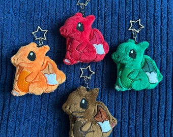 Puede incluir: Cuatro llaveros de dragón de peluche en naranja, rojo, verde y marrón. Cada criatura suave y rellena presenta detalles bordados, grandes ojos negros, alas pequeñas y una punta de cola blanca. Un cierre de metal plateado en forma de estrella está sujeto en la parte superior.