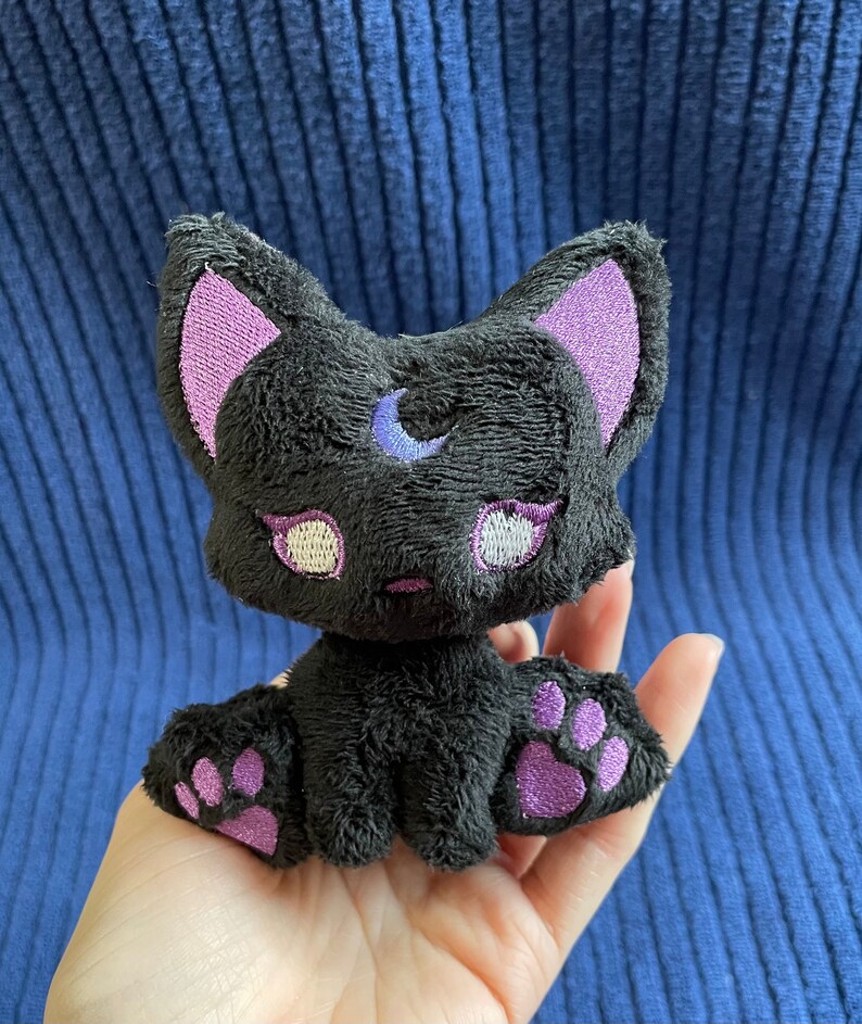 May include: Black plush cat toy with purple embroidered ears, paws, and a crescent moon on its forehead. The cat has white embroidered eyes.