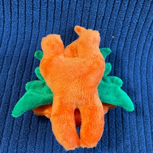 Puede incluir: Juguete de peluche naranja con forma de zanahoria con hojas de peluche verdes. El juguete est&aacute; tumbado de espaldas con las piernas en el aire.