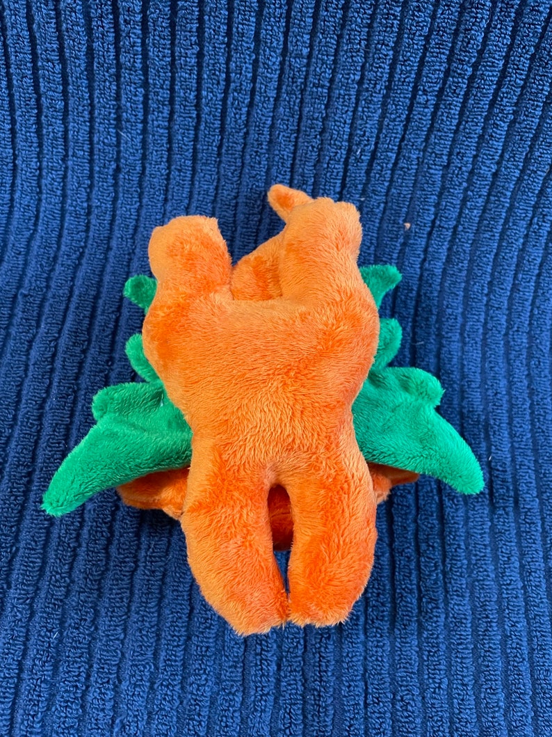 Puede incluir: Juguete de peluche naranja con forma de zanahoria con hojas de peluche verdes. El juguete est&aacute; tumbado de espaldas con las piernas en el aire.