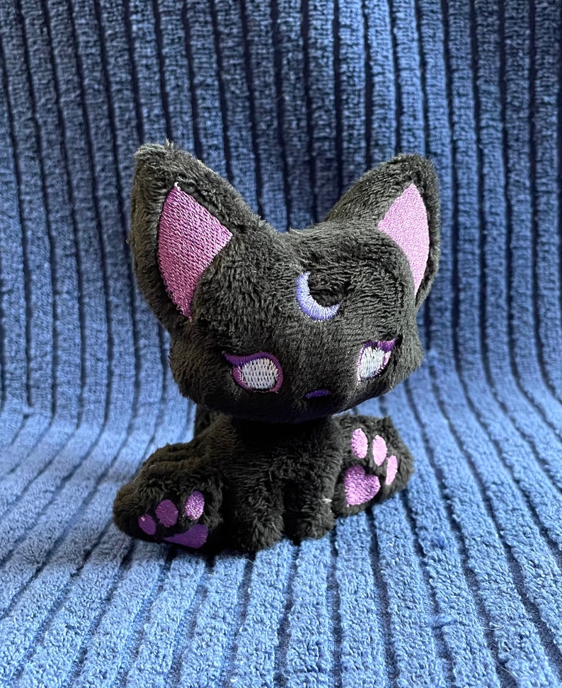 May include: A black plush cat with purple ears and paw pads. The cat has a crescent moon embroidered on its forehead.