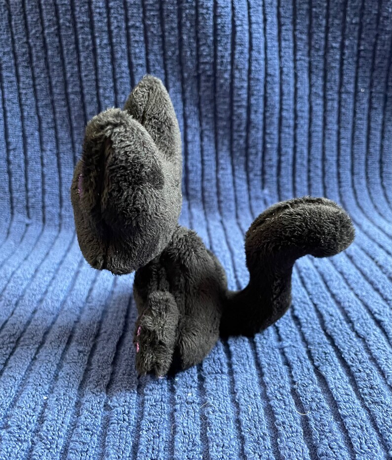 May include: A black plush cat toy with pink accents on the ears and paws. The cat is sitting on a blue textured surface.