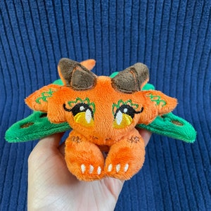 Puede incluir: Un peque&ntilde;o drag&oacute;n de peluche naranja con alas verdes y ojos grandes. El drag&oacute;n est&aacute; sentado sobre sus patas traseras con sus patas delanteras metidas.