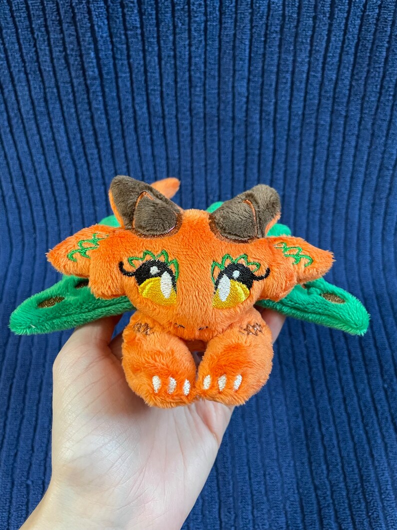Puede incluir: Un peque&ntilde;o drag&oacute;n de peluche naranja con alas verdes y ojos grandes. El drag&oacute;n est&aacute; sentado sobre sus patas traseras con sus patas delanteras metidas.