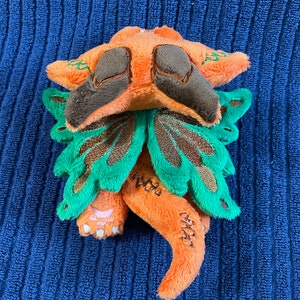 Puede incluir: Un peque&ntilde;o drag&oacute;n de peluche naranja y verde con ojos marrones y alas. El drag&oacute;n est&aacute; enroscado con la cola metida debajo del cuerpo.