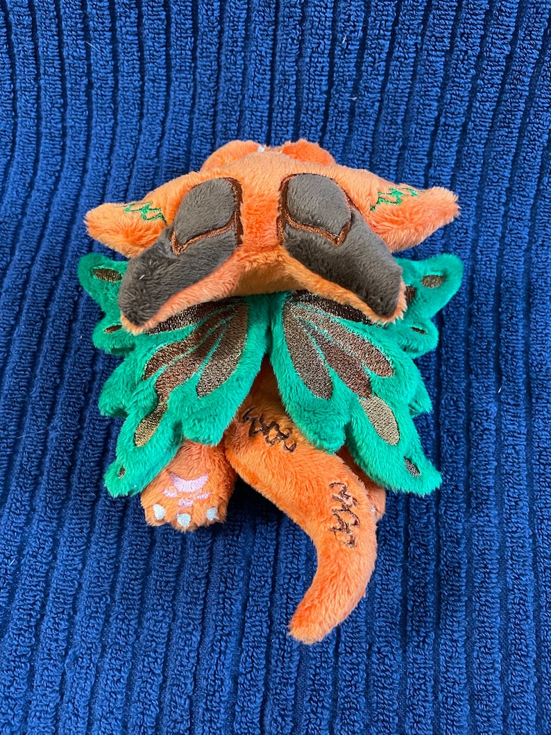 Puede incluir: Un peque&ntilde;o drag&oacute;n de peluche naranja y verde con ojos marrones y alas. El drag&oacute;n est&aacute; enroscado con la cola metida debajo del cuerpo.