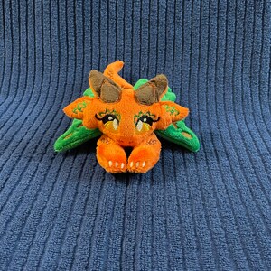 Puede incluir: Un peque&ntilde;o drag&oacute;n de peluche naranja y verde con ojos bordados y una expresi&oacute;n linda. El drag&oacute;n est&aacute; acostado boca abajo con las patas metidas debajo de la barbilla.