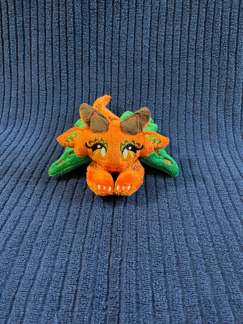 Puede incluir: Un peque&ntilde;o drag&oacute;n de peluche naranja y verde con ojos bordados y una expresi&oacute;n linda. El drag&oacute;n est&aacute; acostado boca abajo con las patas metidas debajo de la barbilla.