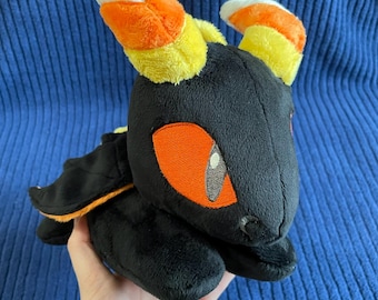 Puede incluir: Juguete de peluche negro con ojos naranjas y cuernos de caramelo. El juguete está acostado de lado con las alas extendidas.